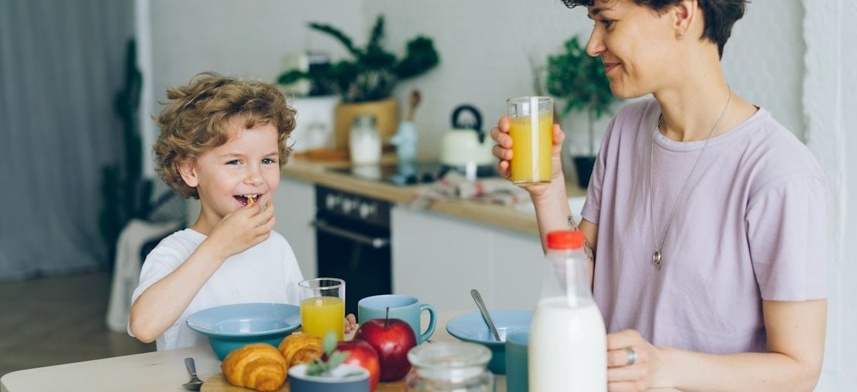 Young woman drinking orange juice while son eating cereal at table in kitchen