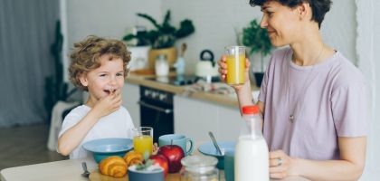 Young woman drinking orange juice while son eating cereal at table in kitchen