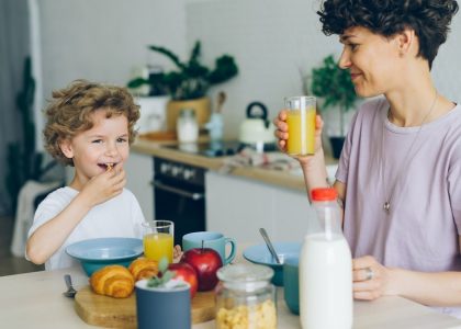 Young woman drinking orange juice while son eating cereal at table in kitchen